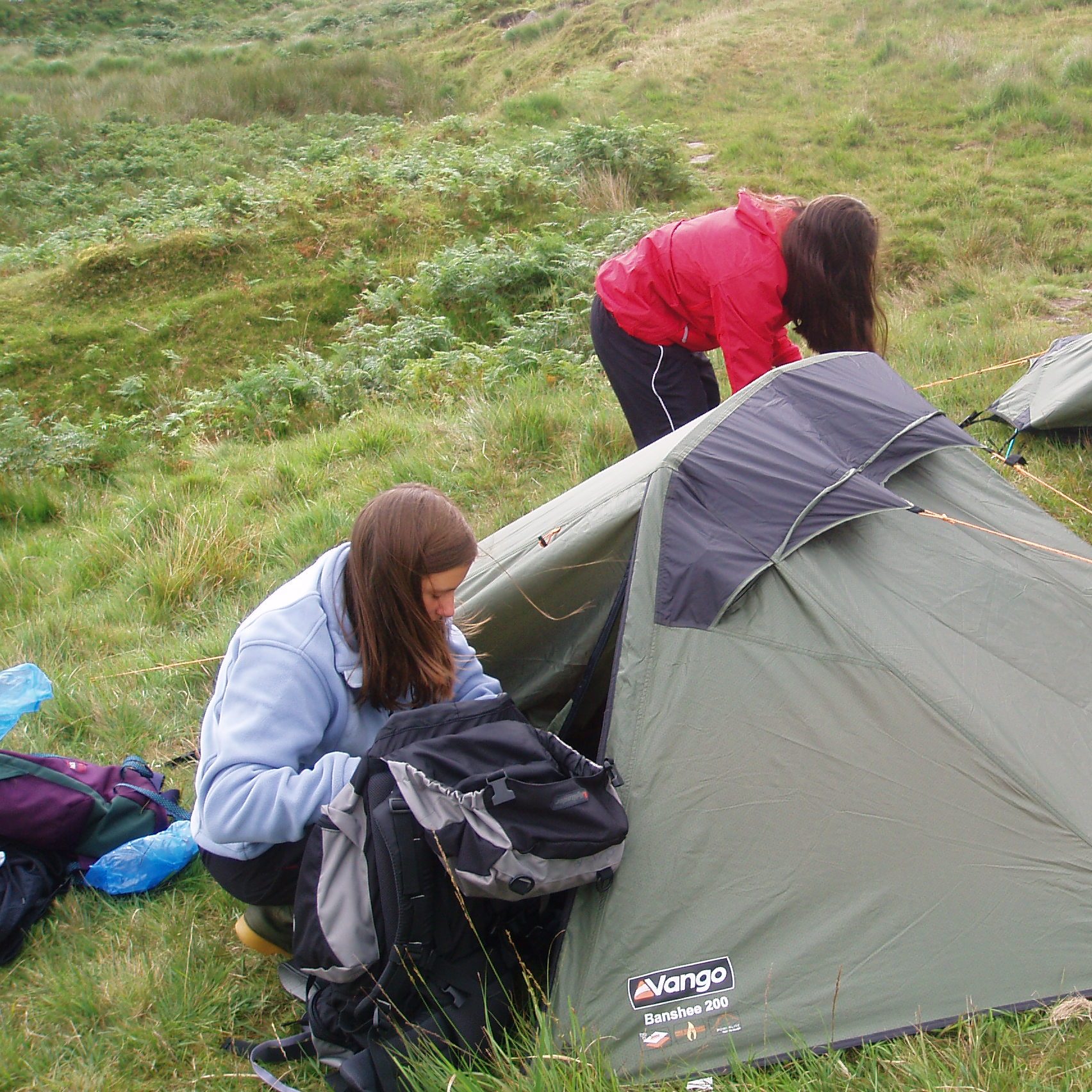 Two children unpacking rucksacks next to a small tent surrounded by bracken and grass