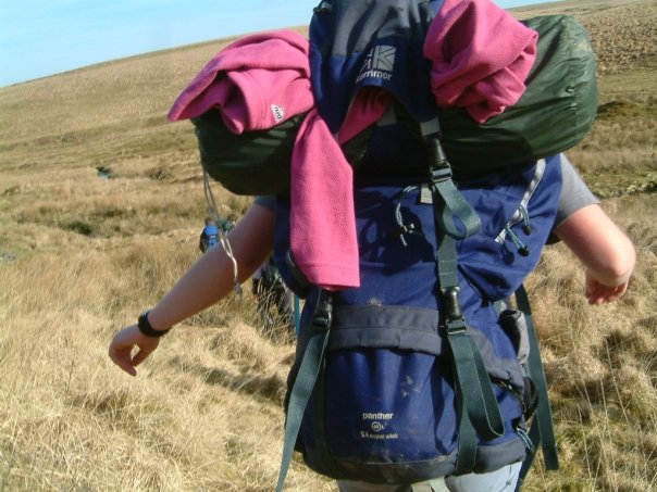 A back view of someone carrying a big rucksack across the moor, with a tent and fleece in the top of the bag